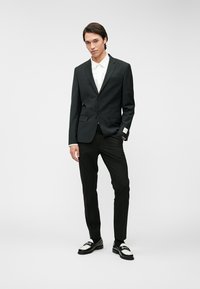 Homem de pé com uma mão no bolso, vestindo um fato preto, camisa branca, calças pretas e mocassins em preto e branco, contra um fundo simples.