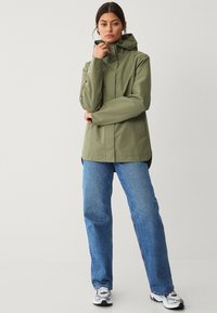 Chubasquero verde oliva impermeable con cuello alto y capucha ajustable, combinado con unos vaqueros azules holgados y deportivas blancas.