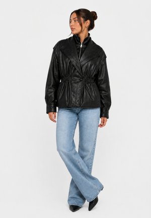 Kvinde iført en sort quiltet jakke med elastisk talje og lyseblå, udvidede jeans, står med krydsede ben og ser til siden.