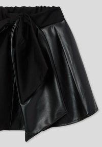 Gonna in ecopelle nera con una fascia larga in vita, caratterizzata da un grande fiocco in tessuto allacciato frontalmente, dettagli plissettati e una silhouette a campana.