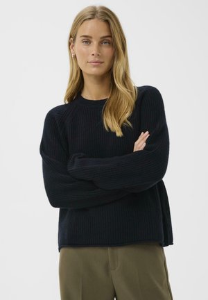 Navy ribbed knit sweater with a crew neck, long sleeves, and a relaxed fit; paired with olive green trousers.