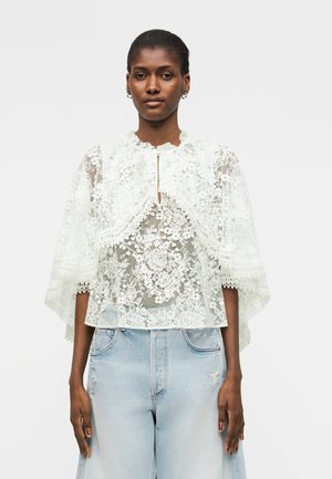 Kobieta w białej kwiecistej koronkowej bluzce z dekoltem typu keyhole i szerokimi rękawami, w połączeniu z jasnoniebieskimi, wysokim stanem jeansami.