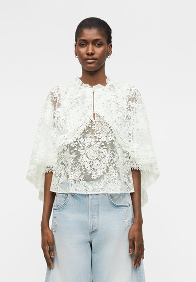 Femme portant un chemisier en dentelle florale blanche avec un col en trou de serrure et des manches larges, associé à un jean taille haute bleu clair.