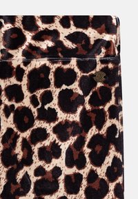 Tkanina s leopardím potiskem, která má měkkou, plyšovou texturu a tmavě hnědé skvrny na béžovém pozadí. Obsahuje malý zlatý logo akcent.