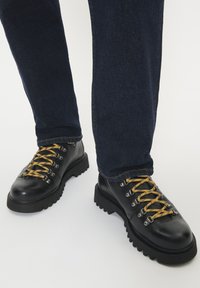 Stivali di pelle nera con suola spessa e lacci gialli indossati con jeans in denim blu scuro su uno sfondo bianco.