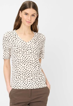 Donna con capelli castani lunghi indossa una maglietta bianca a scollo a V con pois neri e pantaloni marroni, mani in tasca, in piedi contro uno sfondo bianco.