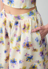 Blumiger Rock mit einem pastellfarbenen Hintergrund, der gelbe, blaue und lila Blumen zeigt. Aus leichtem Stoff mit einem elastischen Bund und Taschen gefertigt.