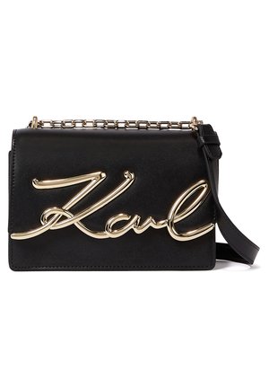 Bolso de hombro rectangular de cuero negro con correa de cadena dorada y grandes letras cursivas doradas en la solapa frontal.