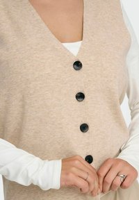 Gilet beige lavorato a maglia con scollatura a V, cinque bottoni neri e texture fine. Indossato sopra una camicia bianca a maniche lunghe. Mani che sistemano il capo.