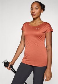 Kortärmad, rundhalsad tröja i rostfärg, tillverkad av ett slätt material. Har en liten vit logotyp och matchas med mörka leggings.