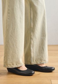 Scarpe a zeppa nere con punta arrotondata e finitura liscia, abbinate a jeans a gamba larga di colore chiaro, esposte su un pavimento in legno.
