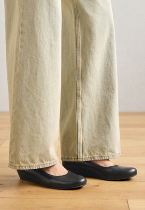 Juodi platforminiai batai su apvaliu nosiu ir lygiu paviršiumi, derinami su šviesių spalvų plačiais džinsais, išdėstyti ant medinės grindų.