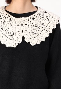 Pull noir avec un grand col blanc en crochet présentant des motifs floraux et circulaires, ajoutant texture et contraste au design.