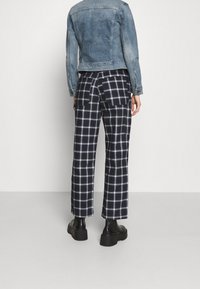 Persona con pantalones de cuadros en blanco y negro, botas de plataforma negras y una chaqueta de mezclilla azul, de pie frente a una pared blanca.
