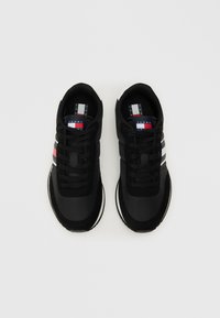 Sapatilhas pretas com um upper texturizado, apresentando riscas brancas e vermelhas. Bico arredondado, sola almofadada e logótipo na língua.