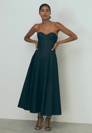 Vrouw met donker haar in een strapless, aansluitende zwarte jurk met zichtbare stiksels, staand met handen in de zij tegen een effen witte achtergrond.
