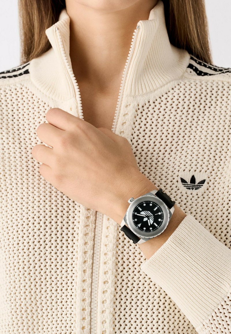 Montre en argent avec un cadran noir et un boîtier rond, dotée d'un bracelet en caoutchouc. Portée sur une main près d'un pull en beige texturé avec un col zippé.