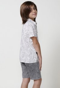 Trumpas rankovėmis baltas marškinis su pilkais lapų raštais, derinamas su pilkomis džinsinio audinio šortais. Marškiniai turi apykaklę su mygtukais ir šoninį ženkliuką.