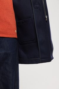 Giacca blu navy in misto lana con chiusura a zip, tasche laterali e un maglione arancione a contrasto sotto, abbinata a jeans scuri.