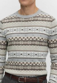 Man in een lichtgrijze, beige en bruine Fair Isle gebreide trui met ronde hals en een bruine gevlochten riem op grijze broek.