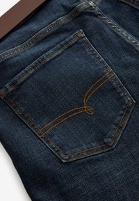 Tylnia kieszeń dżinsowa w ciemnym niebieskim kolorze, z żółtym przeszyciem i unikalnym elementem wzorniczym. Tekstura jest gładka z delikatnym wyblaknięciem.