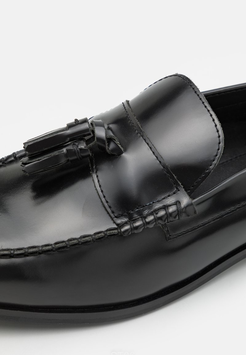 Zapato negro de piel de tipo slip-on con punta redonda, acentos de borlas en la parte frontal y costuras en contraste a lo largo de los bordes. Acabado liso y brillante.
