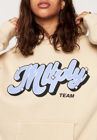 Felpa beige con tasca frontale presenta una grafica audace in nero e azzurro chiaro con il testo "Mltply TEAM," accentuato da motivi a stella.