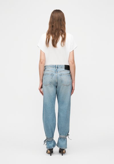 Γυναίκα με μακριά καστανά μαλλιά που φοράει λευκό μπλουζάκι με κοντά μανίκια, ανοιχτόχρωμα γαλάζια τζιν ψηλόμεσα με δεσίματα στον αστράγαλο και μαύρα παπούτσια με ψηλό τακούνι, όρθια.