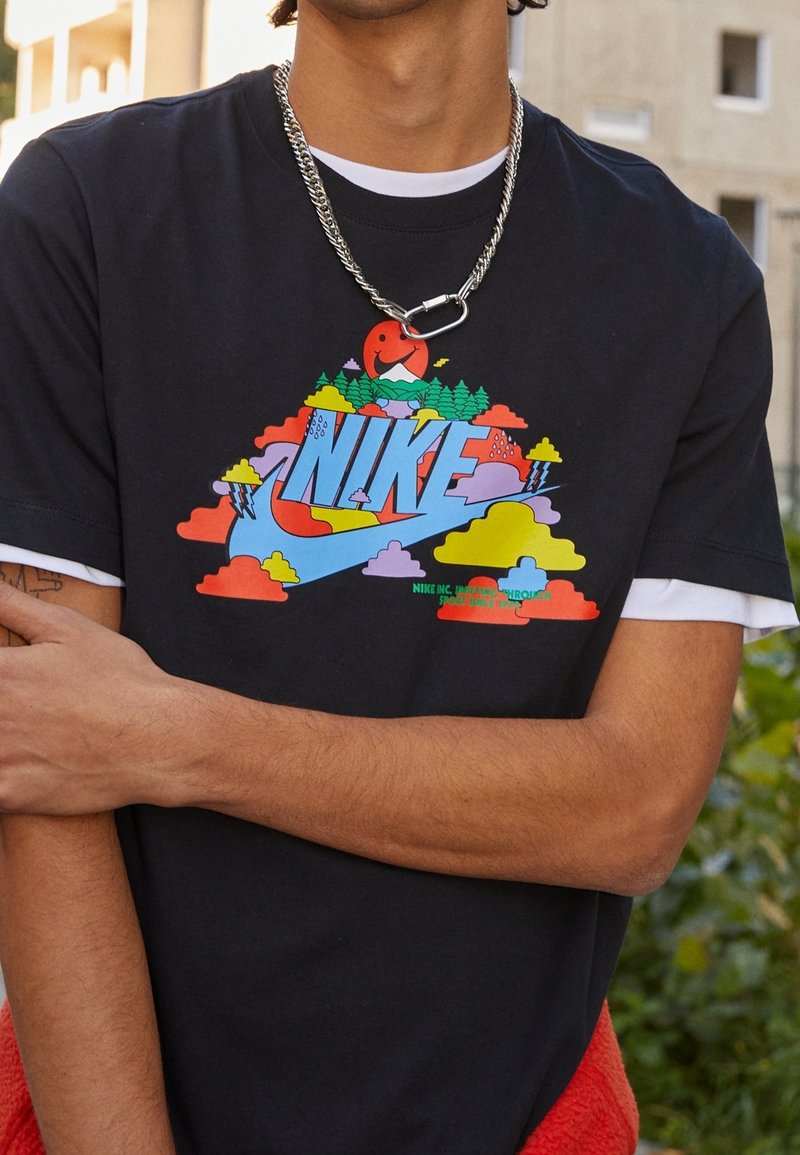 Černé bavlněné tričko s barevným grafickým designem se slovem "NIKE", mraky a stromy. Pod tím je vrstvený bílý výstřih.