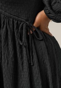 Vestido negro de textura con cintura fruncida, que presenta un detalle de lazo en un lado y mangas largas abullonadas. La tela tiene un patrón sutil.