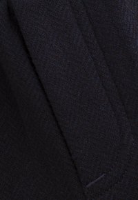 Donkere stof met een subtiel zigzagpatroon en zichtbare naden, die textuur en stikseldetails toont.