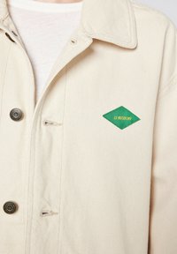 Beige denimjakke med en grøn diamantformet label med "LA MAISON MV" i gul. Metalknapper og krave-design.