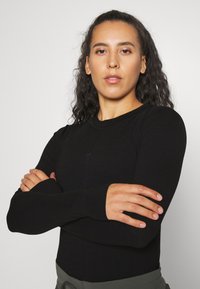 Long-sleeved black top with a fitted design, ribbed texture, and thumbholes at cuffs. Straight neckline and subtle seam detailing.