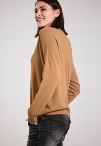 Camel-colored, soft knit sweater with a relaxed fit, ribbed cuffs, and hem, paired with dark gray jeans. Plain textured design.