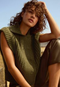 Pull sans manches en tricot vert olive avec un col haut et un motif texturé, associé à un pantalon lisse beige clair.