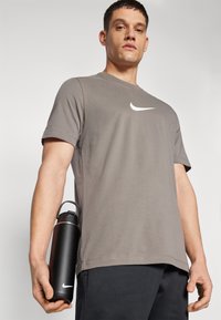 Camiseta gris de manga corta con el logotipo de swoosh de Nike en blanco, combinada con pantalones cortos negros. El modelo sostiene una botella de agua negra con un borde plateado.