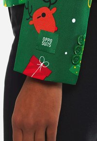Vihreä kangas, jossa on poro- ja lahjakuosiset koristeet, sisältää vihreän ommellun tarran "OPPO SUITS" ja neljä vihreää nappia hiha-mansetissa.