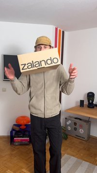 kartonska škatla z logotipom "zalando" v debelih črnih črkah, ki jo drži oseba oblečena v bež flis jakno in črne cargo hlače.