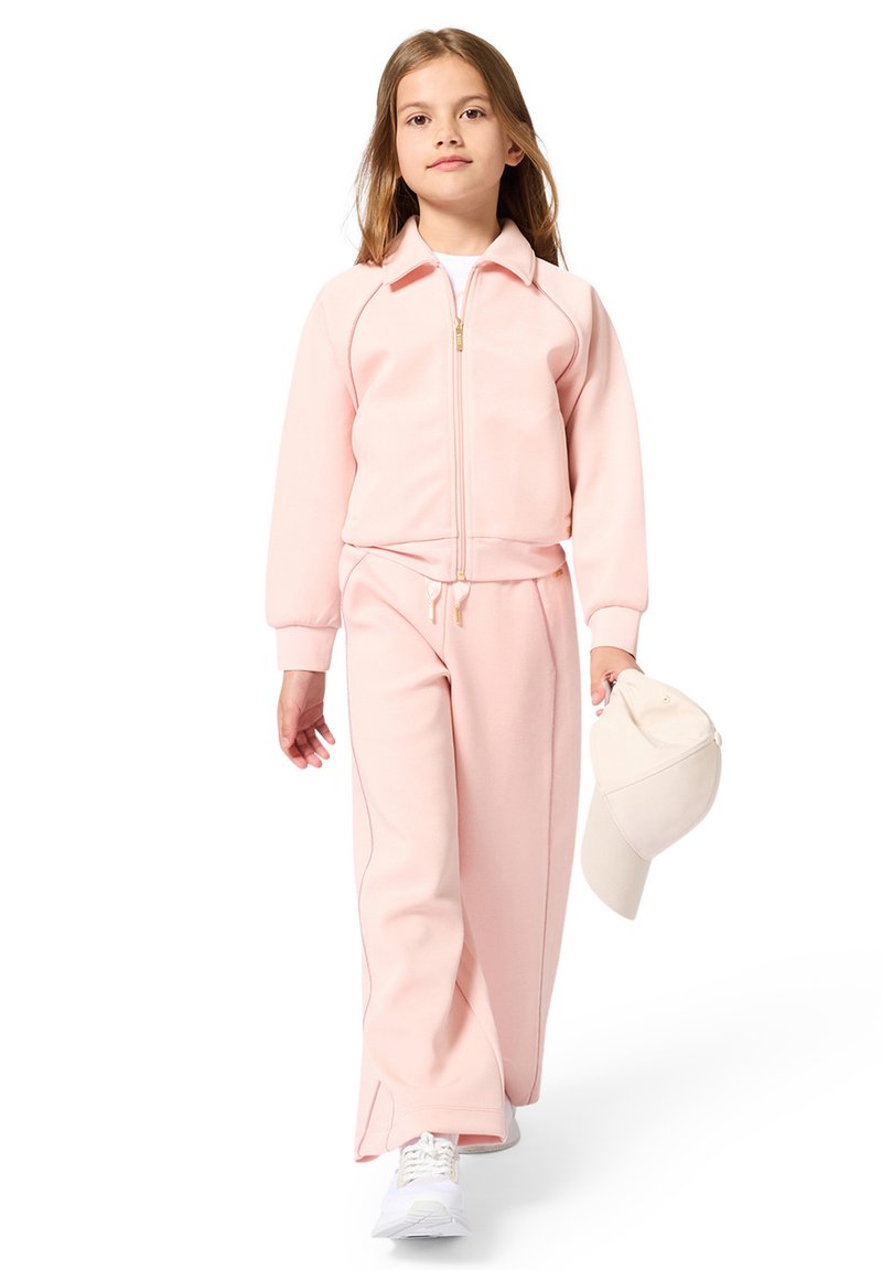 Fille portant une veste zippée rose clair et un pantalon assorti, tenant une casquette blanche dans sa main droite, marchant avec assurance.