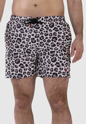 Mann trägt Badeshorts mit Leopardenmuster und schwarzem Kordelzug, gezeigt von der Taille bis zur Mitte des Oberschenkels vor einfarbigem Hintergrund.