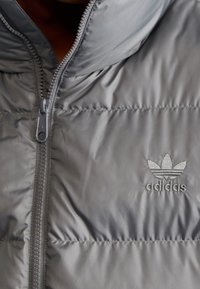 Grå puffjacka i glänsande material, med hög krage, dragkedja och en broderad Adidas-logotyp på vänster sida.