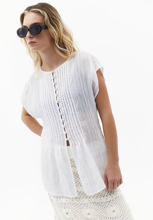 Kobieta w dużych czarnych okularach przeciwsłonecznych, luźnej białej koszuli zapinanej na guziki i białej szydełkowej spódnicy, pozująca na jednolitym tle.