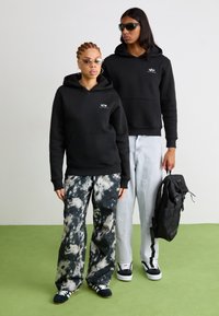 Des sweats à capuche noirs avec des poches à l'avant, associés à des pantalons tie-dye en noir et gris et en denim clair. L'un porte un sac à dos noir et des lunettes de soleil.