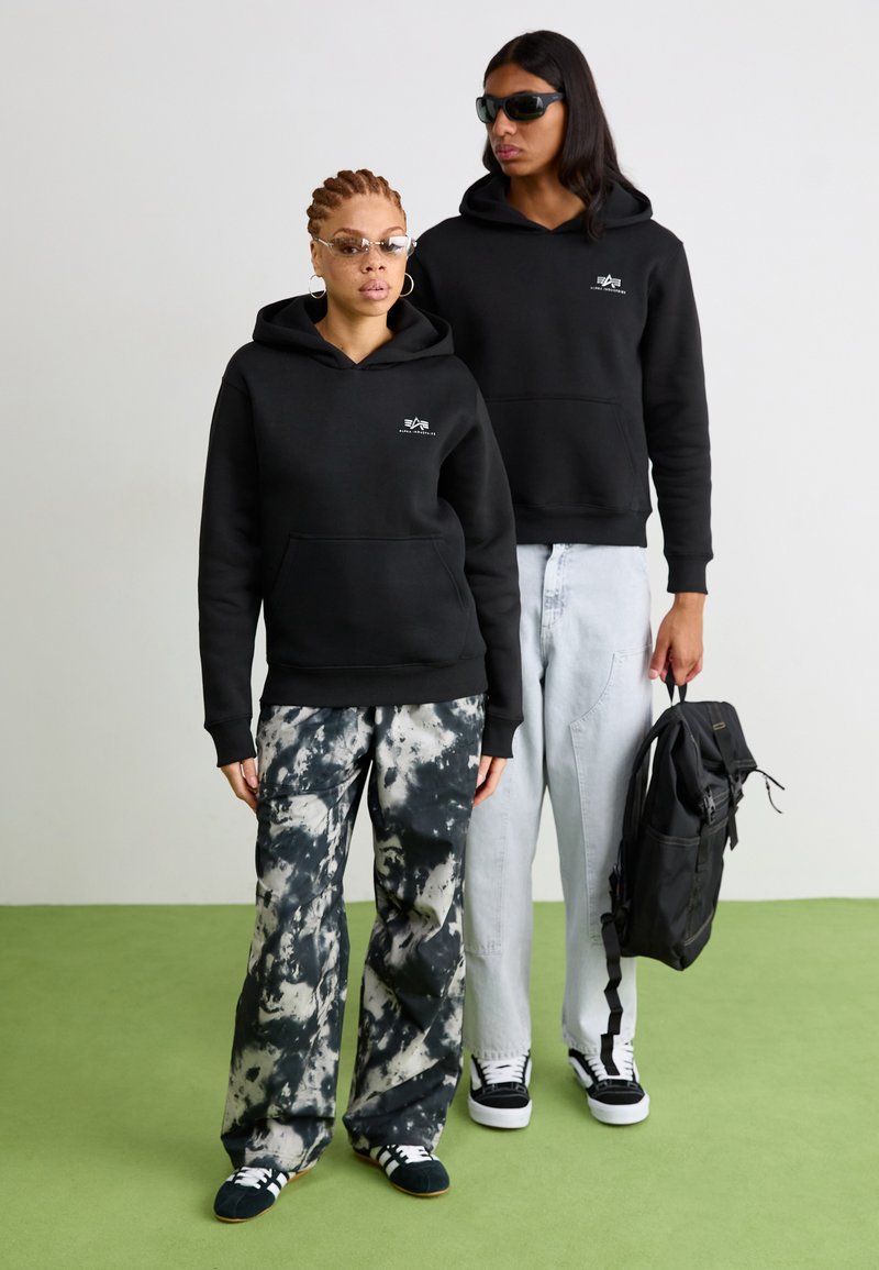 Felpe nere con tasche frontali, abbinate a pantaloni tie-dye in nero e grigio e jeans in denim chiaro. Una persona tiene uno zaino nero e indossa occhiali da sole.