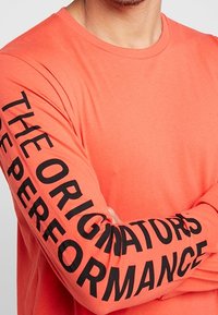 Koszula z długim rękawem, w kolorze koralowym, wykonana z bawełny. Czarny napis wzdłuż lewego rękawa brzmi "TWÓRCY WYKONANIA". Miękka faktura.