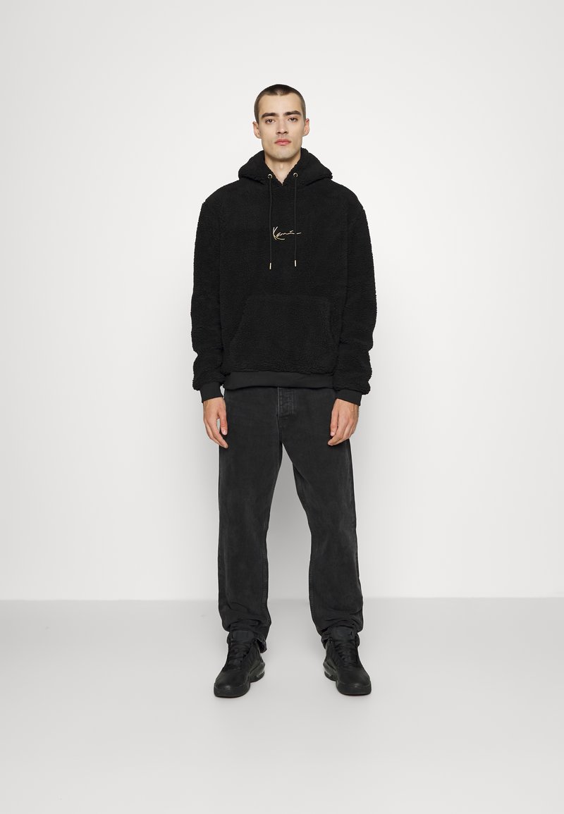 Svart hettegenser i fleece med frontlomme, gulllogo brodering, og svarte jeans. Brukt med svarte joggesko.