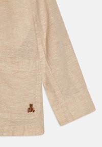 Camicia di lino beige a maniche lunghe, con taschino e dettaglio ricamato di un orso bruno. Tessuto morbido e design rilassato.