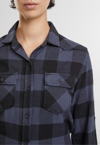 Camicia in flanella con un motivo a quadri navy e nero, chiusura frontale con bottoni e due tasche sul petto con patta e chiusura a bottone.