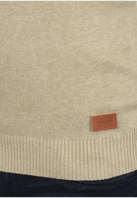 Beige gebreide trui met een gestructureerd oppervlak, voorzien van een geribbelde zoom en een leren label aan de onderlinkerkant.