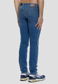 Jeans in denim blu con taglio slim, dotati di etichetta in vita, tasche posteriori e cuciture a contrasto. Indossati con sneakers blu e bianche.